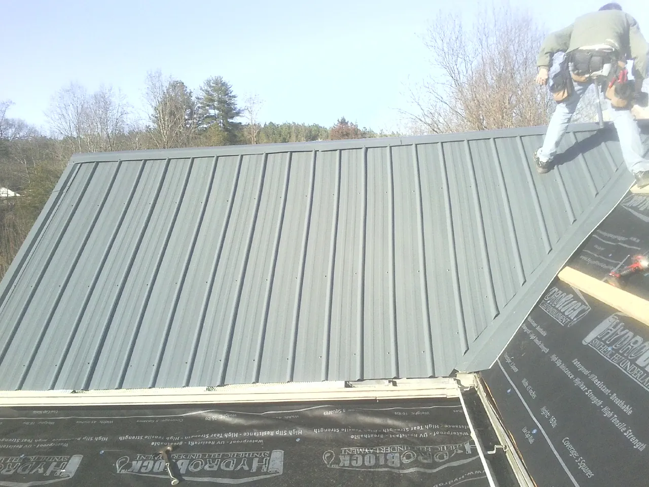 Crew installing dark green standing seam panels over synthetic underlayment in Wardensville