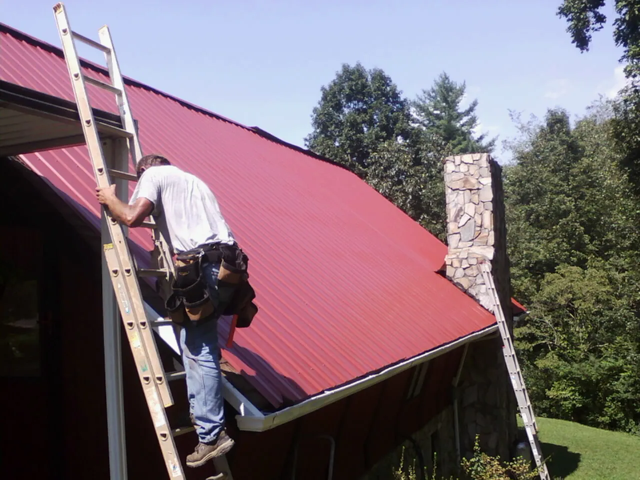 Roofer on a ladder beside a red metal roof and stone chimney — Soffit & Fascia Repair by Roofing Brothers in Wardensville